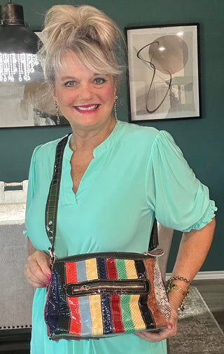 A woman with short blonde hair wearing a mint green dress holds a colorful striped crossbody bag with black trim in an indoor setting.