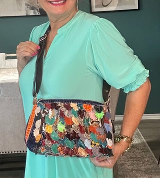 A woman with blonde hair wearing a mint green dress holds a colorful, patterned crossbody bag with a black strap in an indoor setting with a dark green wall.