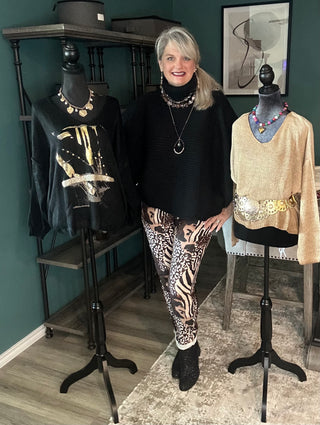 A woman stands between two dress forms wearing and displaying fashion pieces in a boutique setting. She wears a black turtleneck and leopard-print pants, while the mannequins display a black graphic top and a beige patterned sweater.