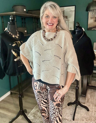 A smiling woman with blonde hair stands in a boutique wearing a cream-colored, textured knit poncho-style top and brown-and-black animal print pants. Mannequins wearing black tops are visible in the background against a dark green wall.