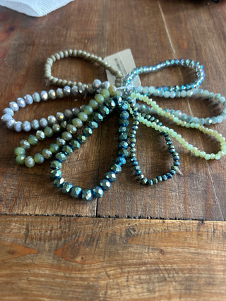 A set of faceted bead strands in assorted colors including blue, green, gray, and beige, arranged on a rustic wooden surface with a small paper tag attached.