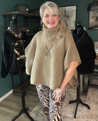 A smiling woman with blonde hair stands indoors wearing a ribbed beige turtleneck poncho sweater and patterned leggings. Two black mannequins displaying clothing are visible behind her against a dark green wall.