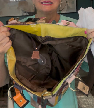 A woman holding open a multicolor tote bag to show its interior, which features a bright yellow-green fabric lining on the inside and a dark brown lining on the outside. She is wearing a turquoise top in a home setting with a dark teal wall and a hanging lamp in the background.