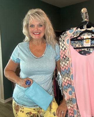 A smiling blonde woman stands indoors holding a light blue clutch, next to a rack of clothing showing a pink tank top and a multicolor patterned cardigan. She is wearing a light blue t-shirt and yellow floral pants.