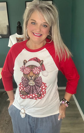 A woman wearing a red and white raglan baseball t-shirt with a festive graphic of a leopard wearing a Santa hat and candy cane eye mask, paired with blue jeans in an indoor setting.