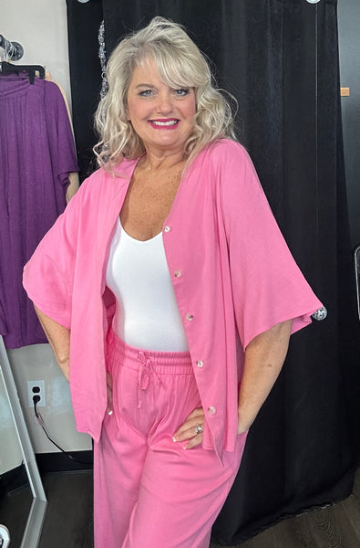 Woman wearing a pink outfit with a white top and pants, standing against a black curtain.