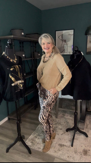 A woman with blonde hair stands in a room wearing a beige long-sleeve top and zebra-print pants, posing between two black dress mannequins displaying clothing.