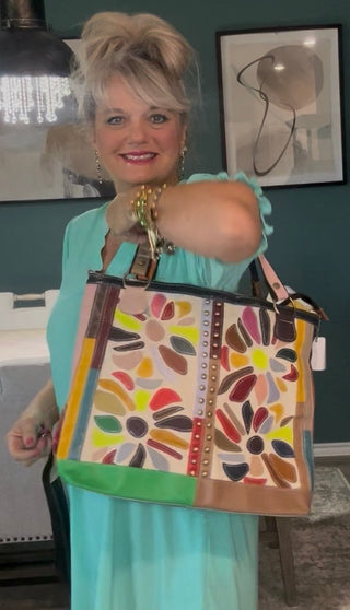 A woman in a mint green dress holds up a large, colorful tote bag with a patchwork floral pattern in shades of orange, yellow, green, red, and brown, set against a dining room background.