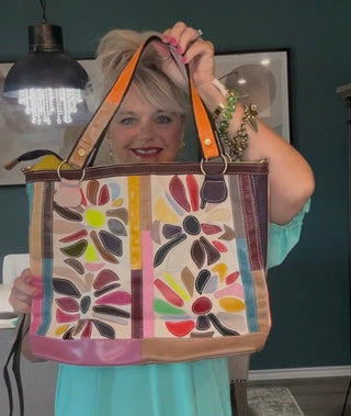A woman in a mint green dress holds up a large canvas tote bag with colorful floral patterns and orange shoulder straps in an indoor room setting.