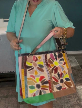 A woman wearing a mint green dress holds up a large, colorful patchwork tote bag with a floral pattern in shades of orange, yellow, green, brown, and pink. The setting is a modern room with a dark teal wall, framed abstract art, and a gray dining table in the background.