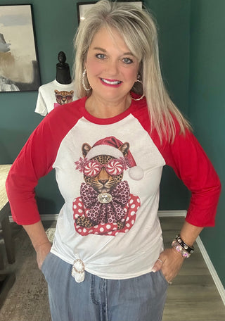 A woman wearing a white and red raglan baseball t-shirt with a festive leopard graphic on the front, posing in an indoor setting with a dark green wall.