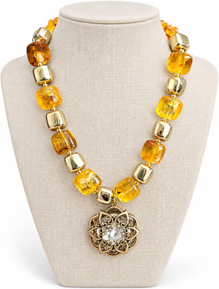 A chunky statement necklace featuring translucent amber-yellow glass beads and polished gold-toned spacer beads, with a central gold-toned filigree medallion holding a clear faceted stone, displayed on a beige fabric jewelry bust.