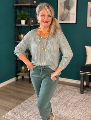 A woman posing in a ribbed sage green long-sleeve top and matching sage green drawstring pants, accessorized with layered gold necklaces, in a styled room with a dark teal wall.