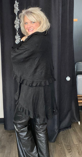 Person wearing a black outfit with ruffled details against a dark curtain.