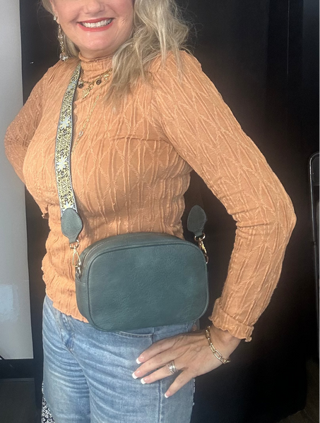 Woman wearing a tan sweater and blue jeans with a green crossbody bag indoors.