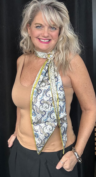 Woman wearing a beige tank top and black pants with a patterned scarf around her neck, standing in a room with black curtains and clothing on a rack.