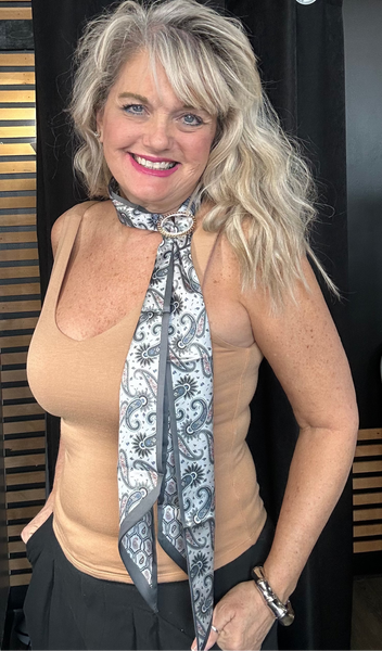 Woman wearing a beige tank top and black skirt with a patterned scarf around her neck, standing in a room with clothing items in the background.