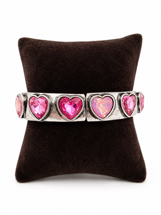 A silver-toned cuff bracelet featuring five heart-shaped pink gemstones, displayed on a dark brown velvet jewelry pillow against a white background.