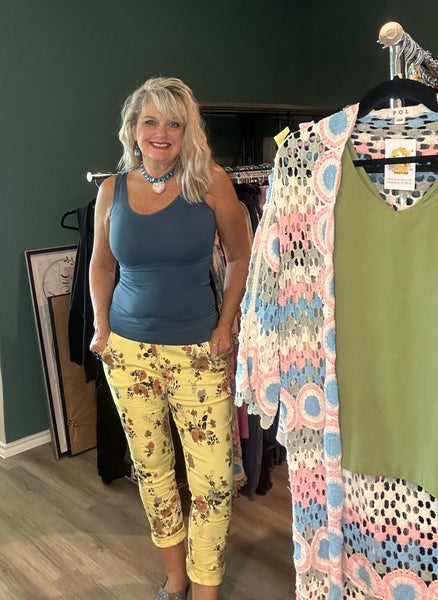 A woman stands in a clothing boutique wearing a slate blue tank top and yellow floral capri pants. To her right, a hanger displays a matching green tank top with a pink, blue, and white crochet patterned cover-up.