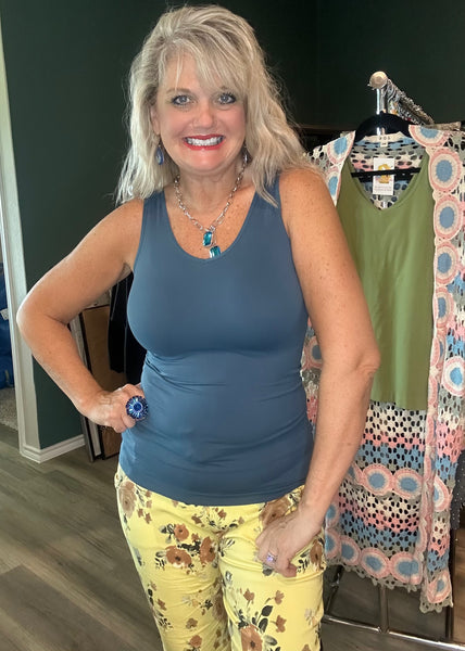 A smiling blonde woman wearing a slate blue tank top and yellow floral pants, standing in front of a clothing rack in an indoor boutique setting.