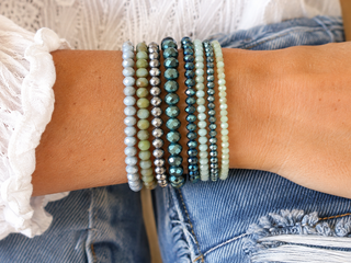 A close-up of a person's wrist wearing a stack of nine beaded stretch bracelets in various colors and patterns, paired with blue jeans and a white lace top.