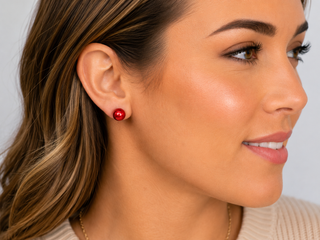 A close-up side profile of a woman wearing a small, round, glossy red stud earring on her earlobe.