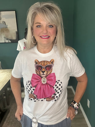 A woman with blonde hair wearing a white graphic t-shirt featuring a leopard wearing sunglasses and a large pink bow, posed in an indoor room with a dark green wall.