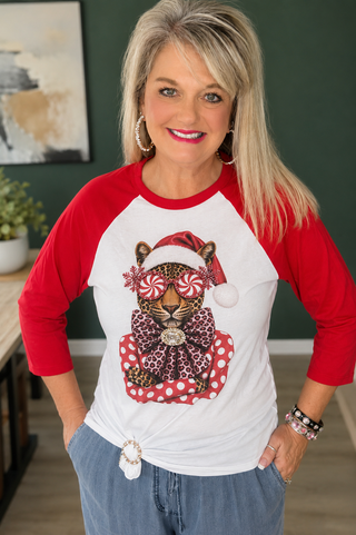 A woman wearing a red and white raglan baseball t-shirt with a festive leopard graphic on the front, paired with blue jeans, standing in an indoor setting.