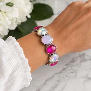 A close-up of a person's wrist wearing a silver-toned bracelet with five oval cabochon gemstones in pink, white, and iridescent colors, styled with a white sleeve and white flowers in the background.