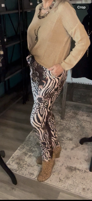 A person wearing a beige long-sleeve top and brown-and-white zebra-print pants with leopard-print ankle boots, standing indoors on a patterned rug.