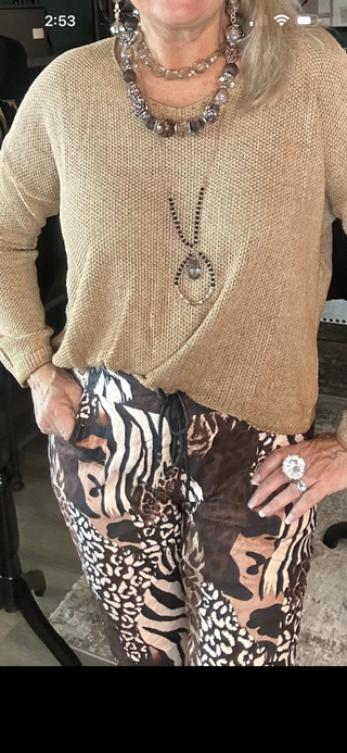A woman wearing a beige knit wrap-style sweater and brown, black, and beige animal-print pants, accessorized with layered necklaces and a ring.