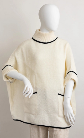 Cream knit poncho sweater with black trim on the mock neck, sleeve cuffs, and hem, displayed on a white mannequin against a light gray background.