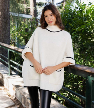 A woman wearing a cream-colored poncho sweater with black trim, standing outdoors on a stone path next to a green railing with trees in the background.
